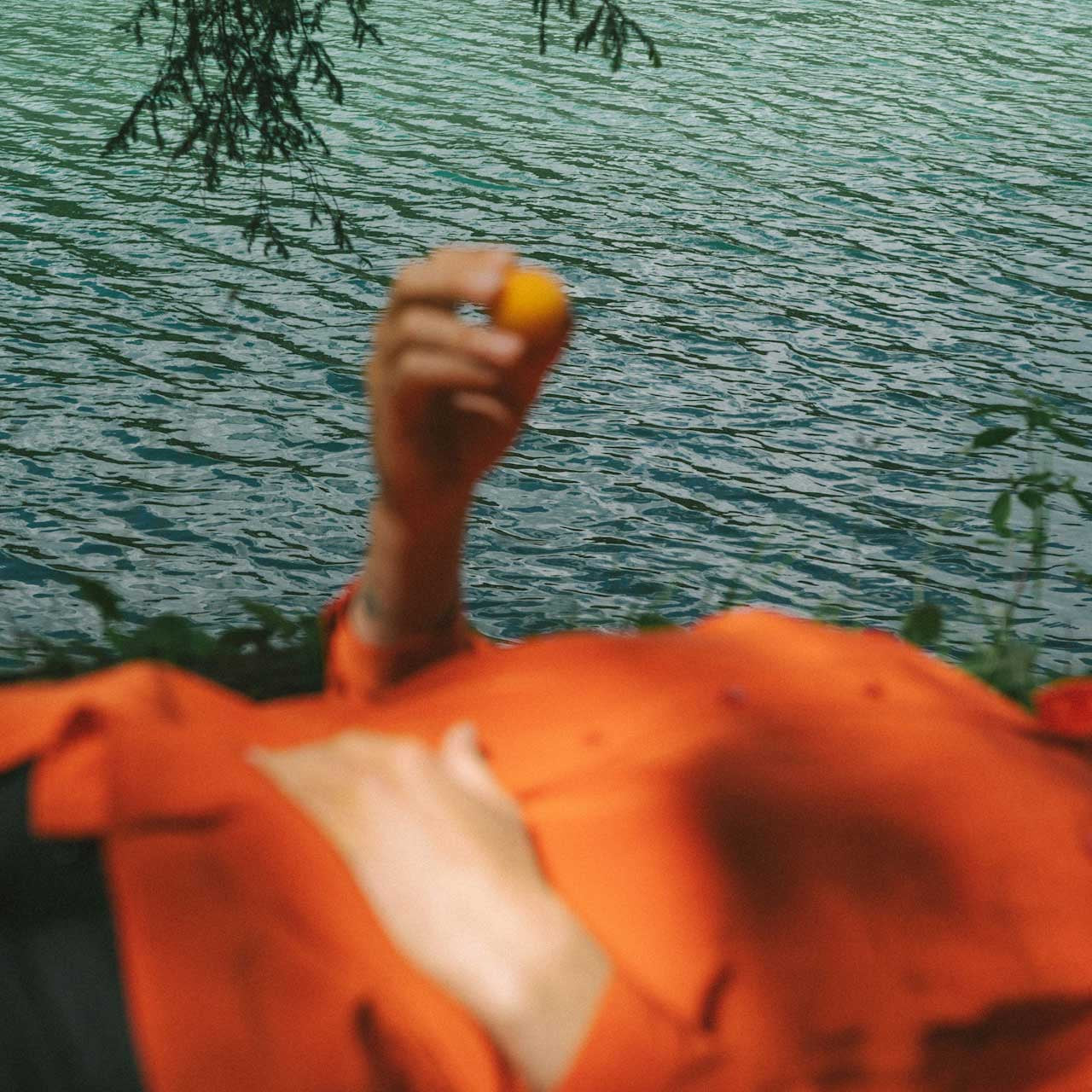 Person in an orange shirt holding a tangerine by a body of water